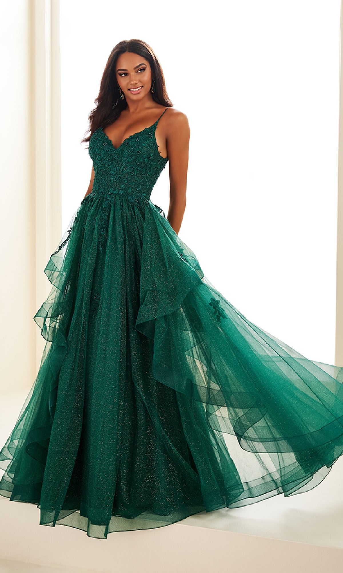 A woman stands indoors wearing the Ellie Wilde EW36085, a floor-length emerald green prom dress with spaghetti straps, a lace embroidered corset bodice, and a layered tulle skirt.